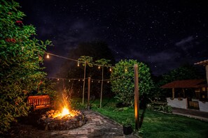 A nighttime outdoor setting with a stone fire pit, string lights, and a wooden bench. - Hotel Amancay - Pool and Jacuzzi  (El Progresso)