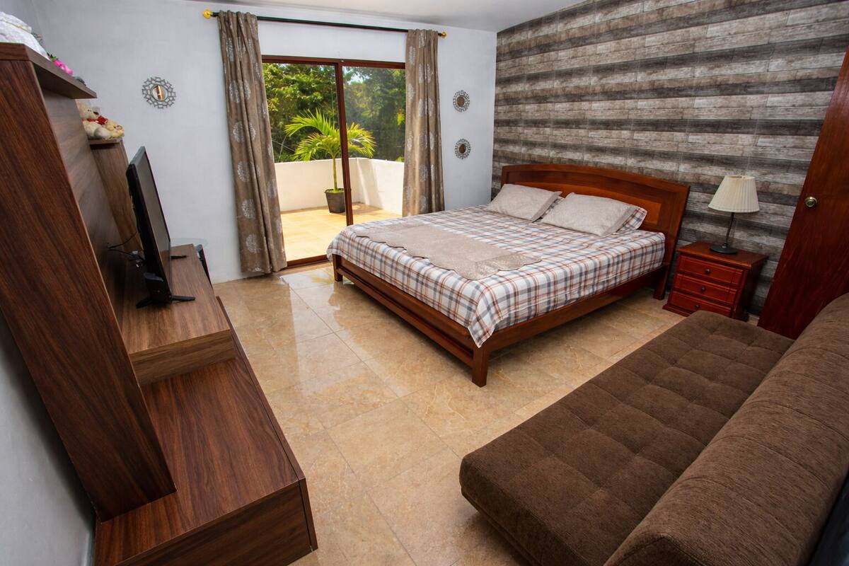 A bedroom with a bed, a sofa, a TV, a nightstand, and a sliding glass door to a balcony.