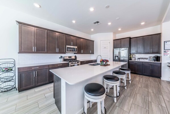 A modern kitchen with dark wood cabinets, stainless steel appliances, a central island with stools, and a white backsplash. - Peaceful 3-bedroom house in Tolleson perfect for relaxing stays (Tolleson)