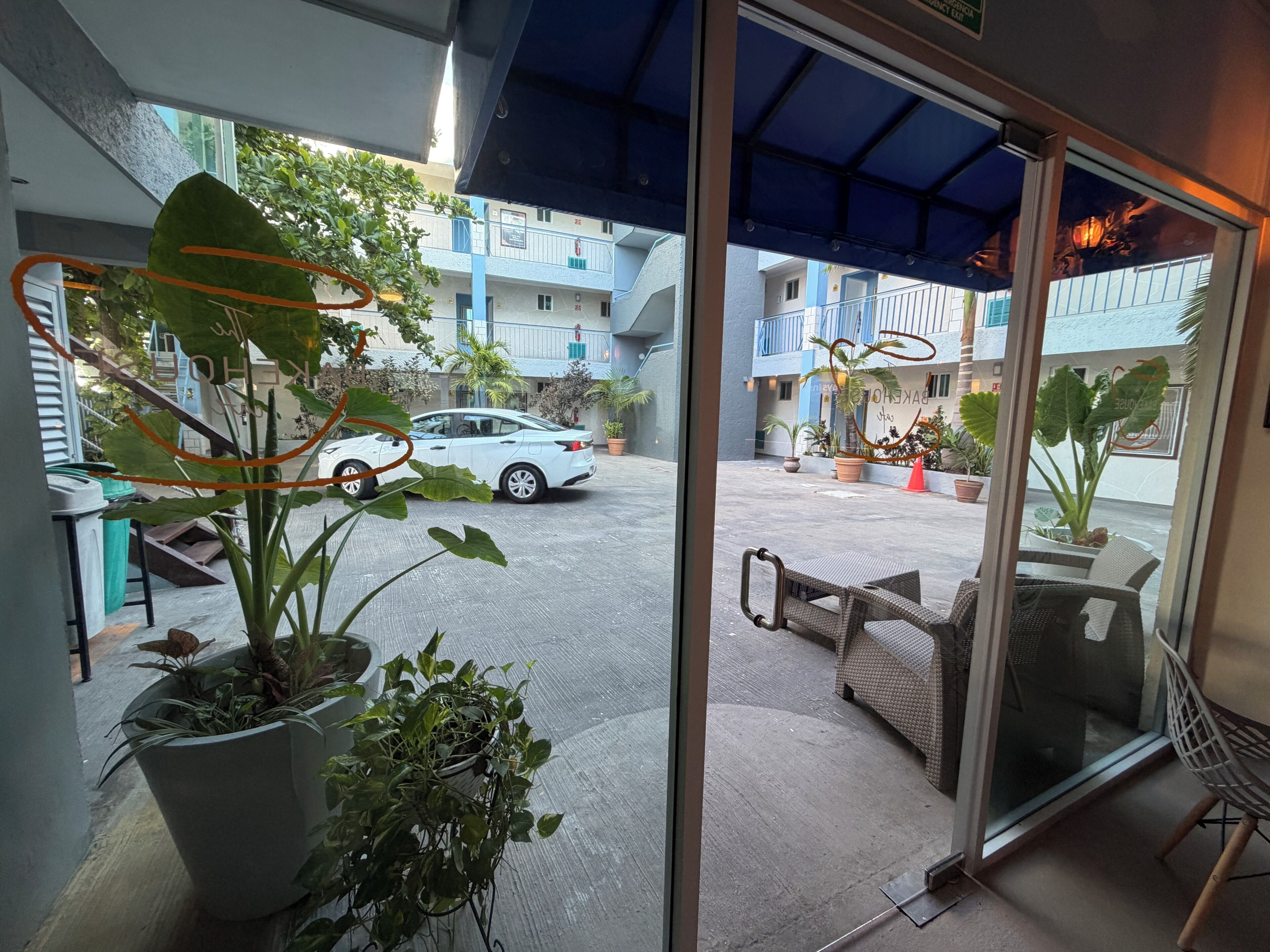 Vista desde el interior de un edificio hacia un patio con un coche blanco estacionado, plantas en macetas y mobiliario exterior.