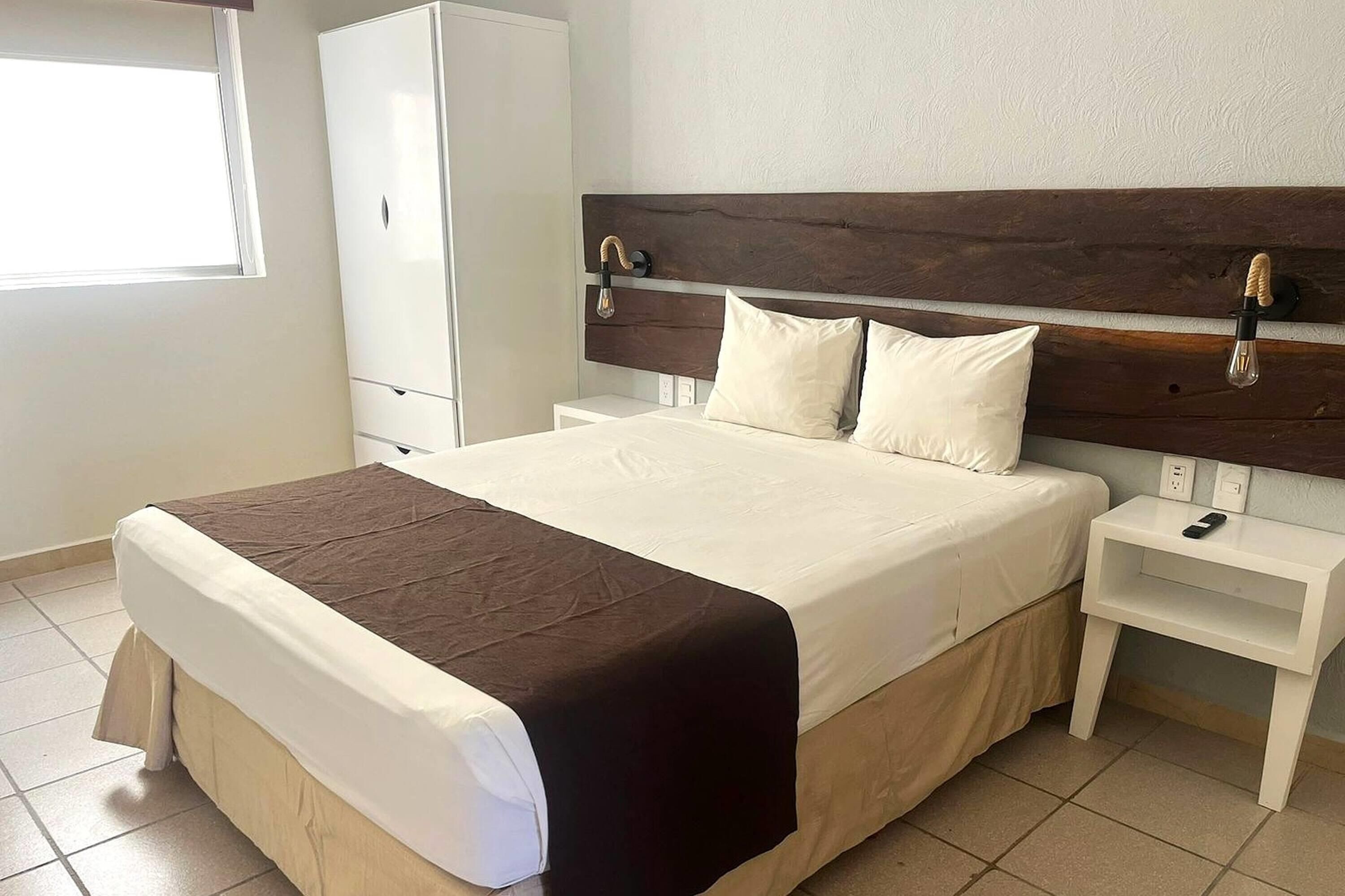 A bed with a wooden headboard, white bedding, a bedside table with a remote control, and a wardrobe.