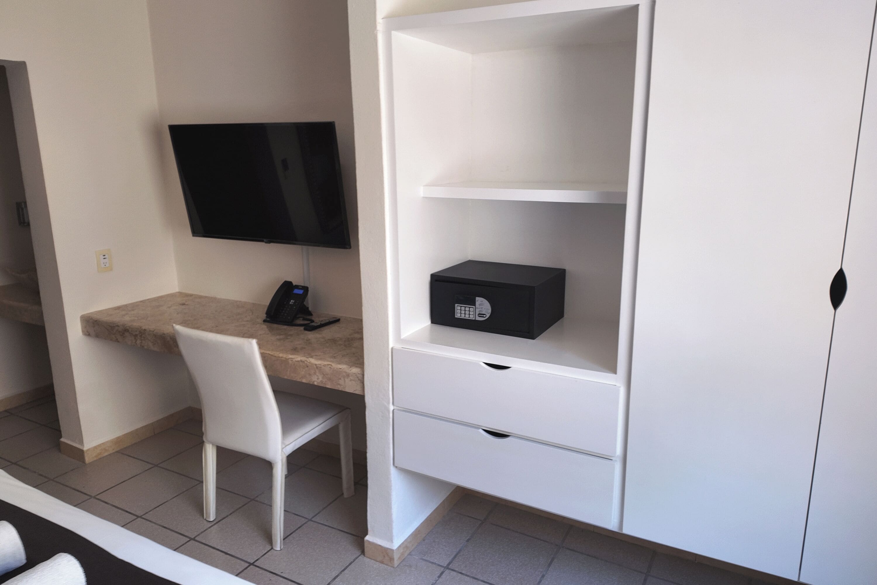 Una habitación moderna con televisión de pantalla plana en la pared, un escritorio con silla y un mueble blanco con cajones.