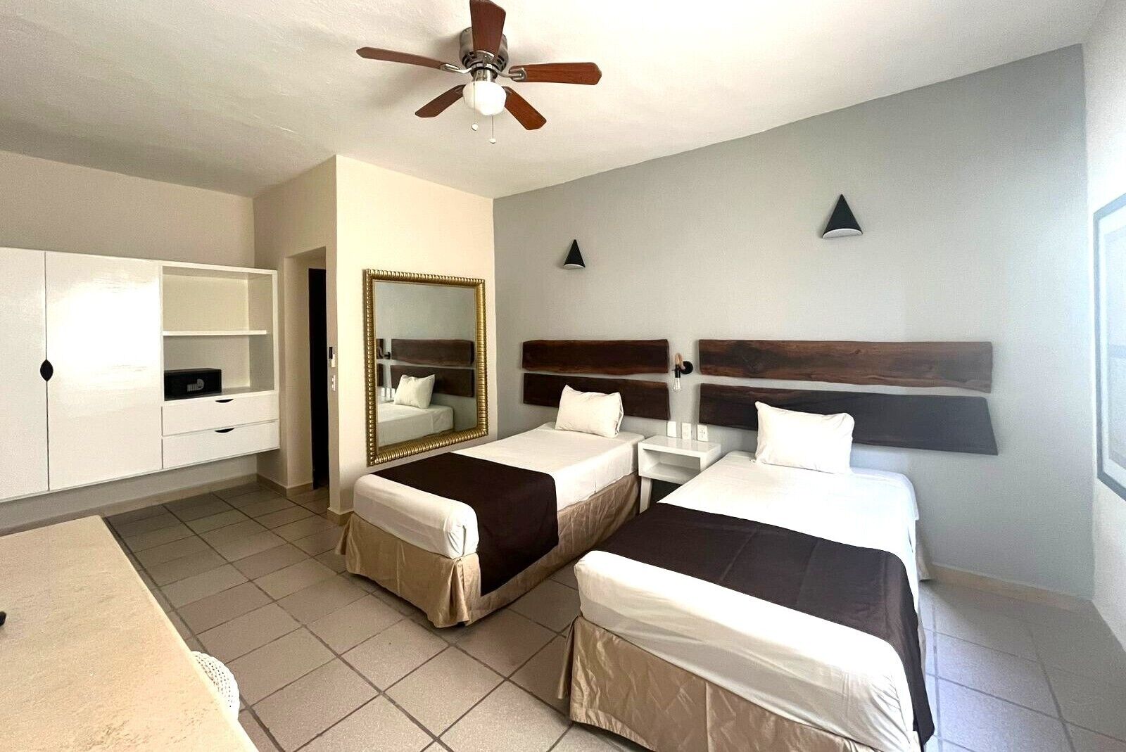 A hotel room with two beds, a wardrobe, a mirror, a ceiling fan, and a small table.