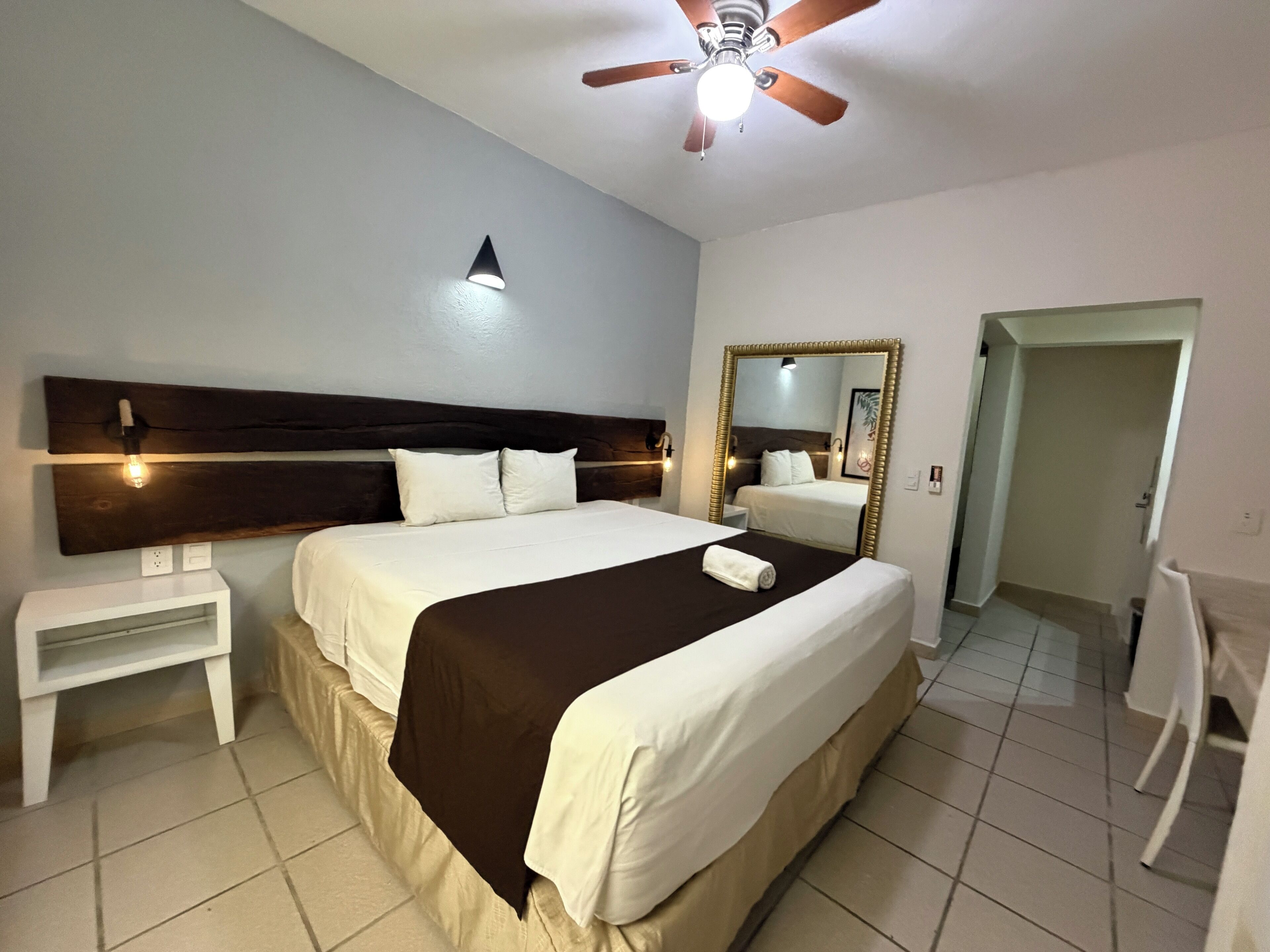 A hotel room with a large bed, a bedside table, a ceiling fan, a wall-mounted light, and a mirror.