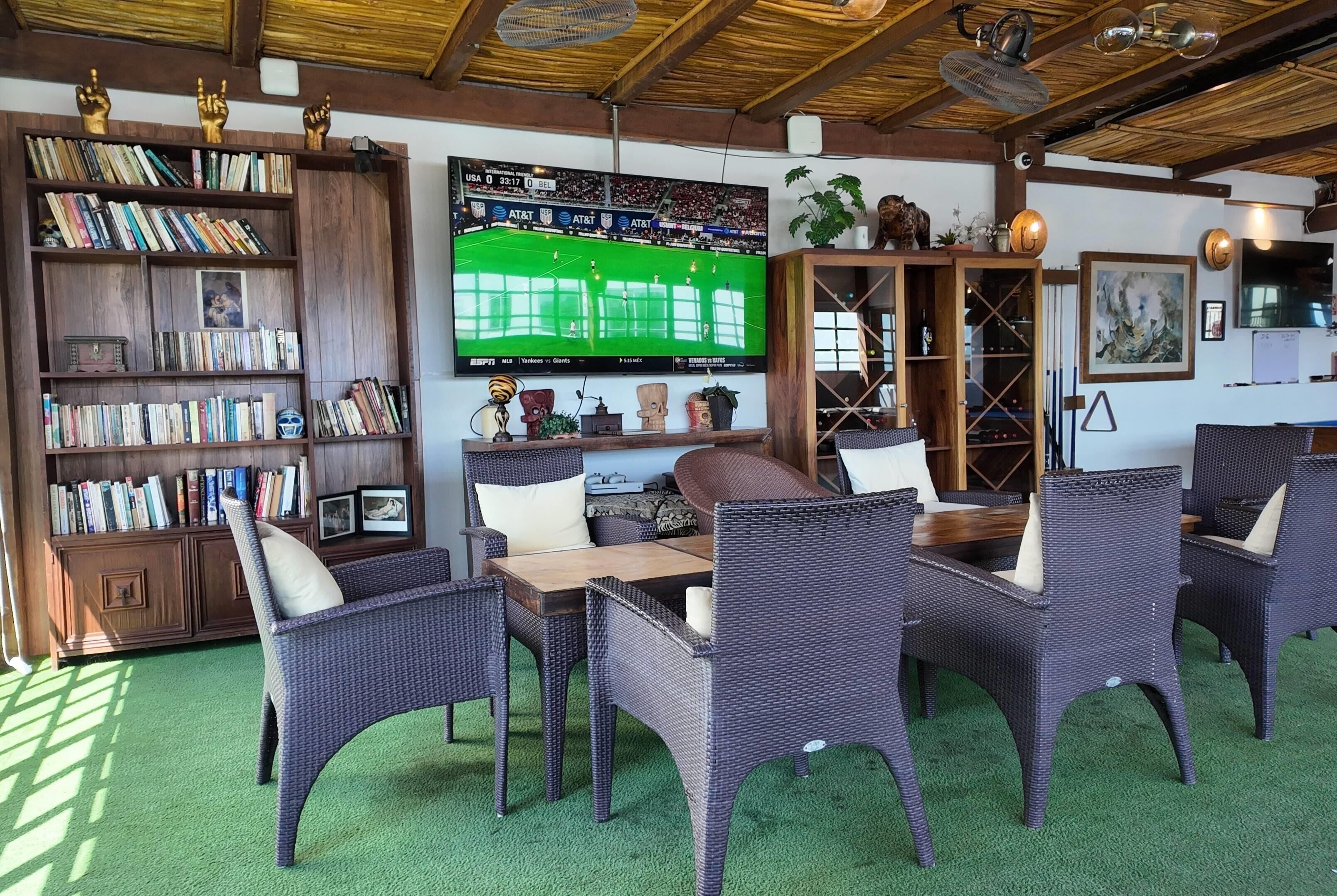 A room with a large flat-screen TV, wicker furniture, a bookshelf, and a ceiling fan.