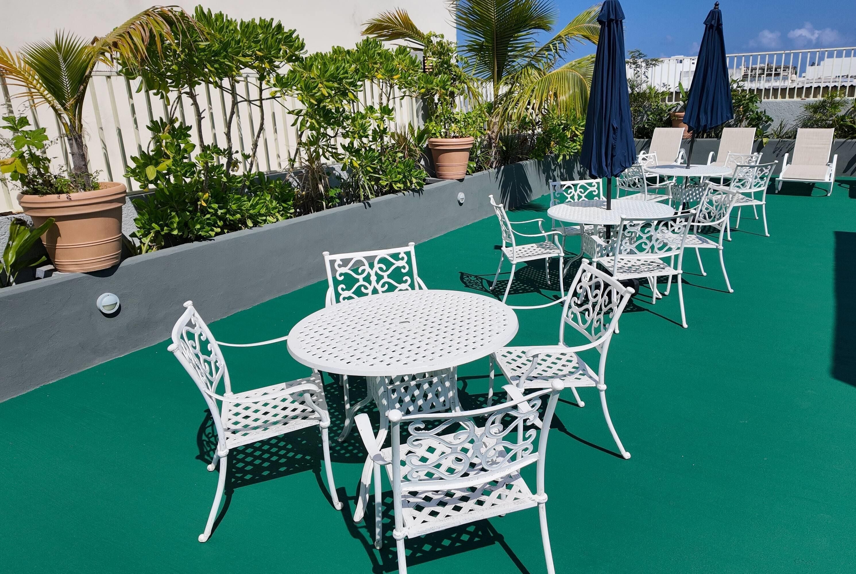 Outdoor patio seating with white metal chairs and tables, surrounded by potted plants and umbrellas.
