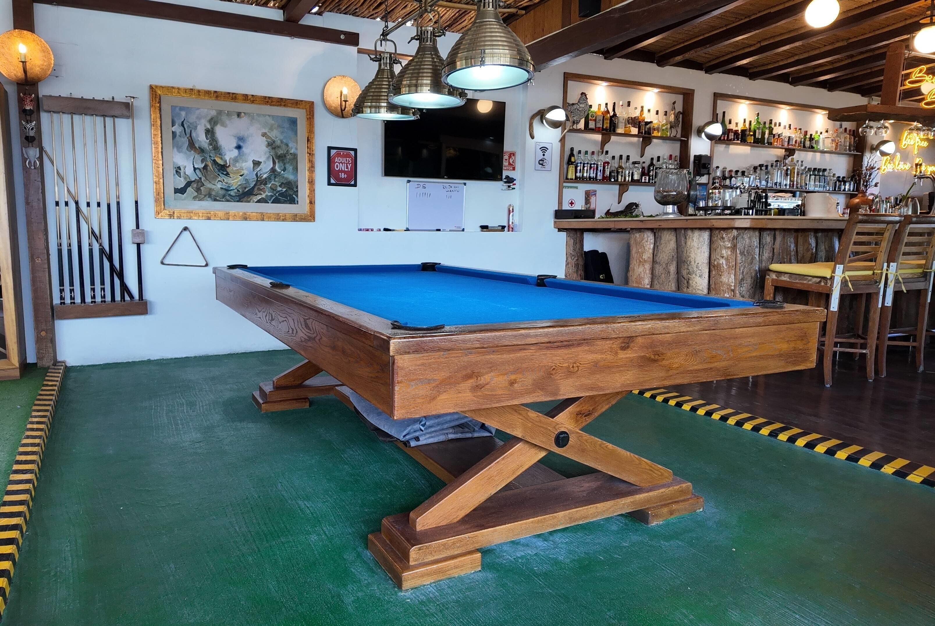 A pool table in a bar area with wooden beams, a painting, and a bar stocked with bottles.