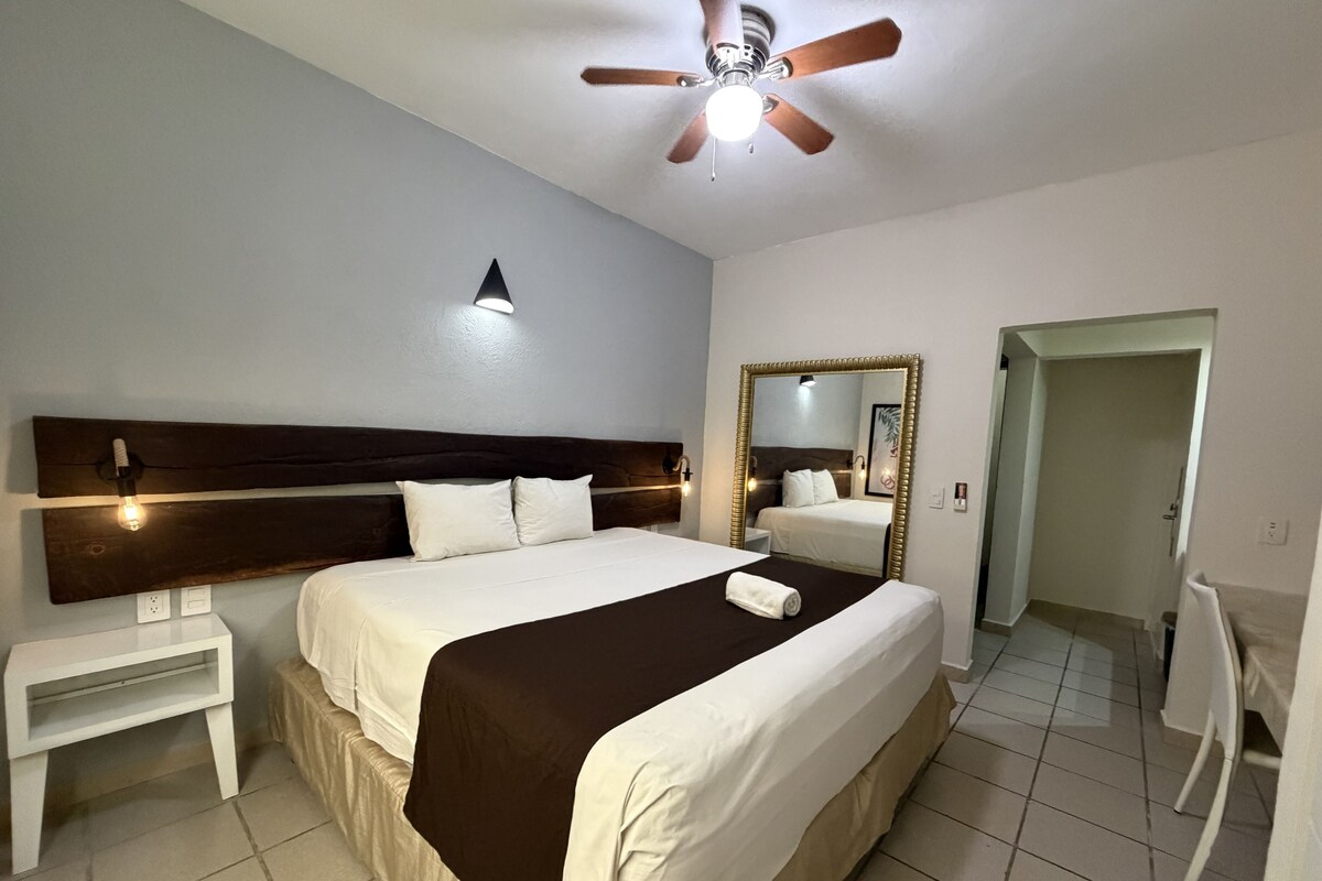 Hotelkamer met een groot bed, een plafondventilator, een nachtkastje, een spiegel en een deur naar een andere kamer.