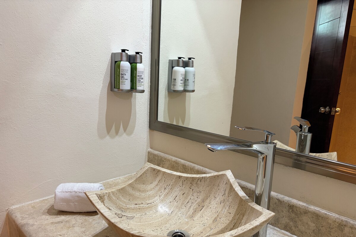A bathroom with a beige stone sink, a mirror, and toiletries on a wall-mounted holder.
