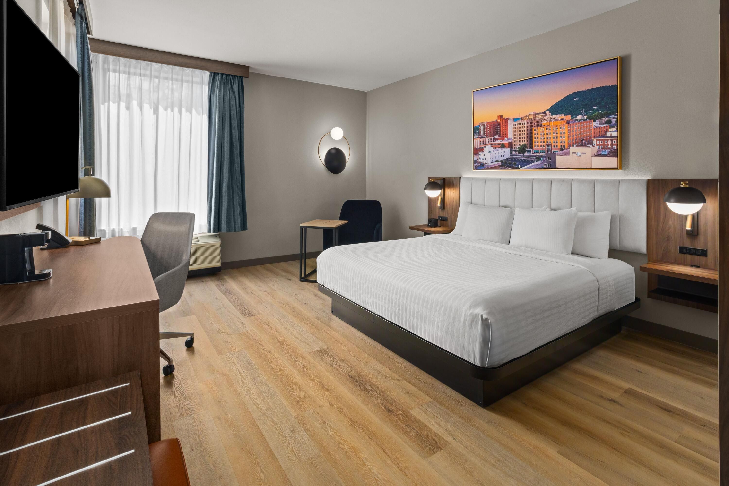 A hotel room with a bed, desk, chair, and a cityscape painting.