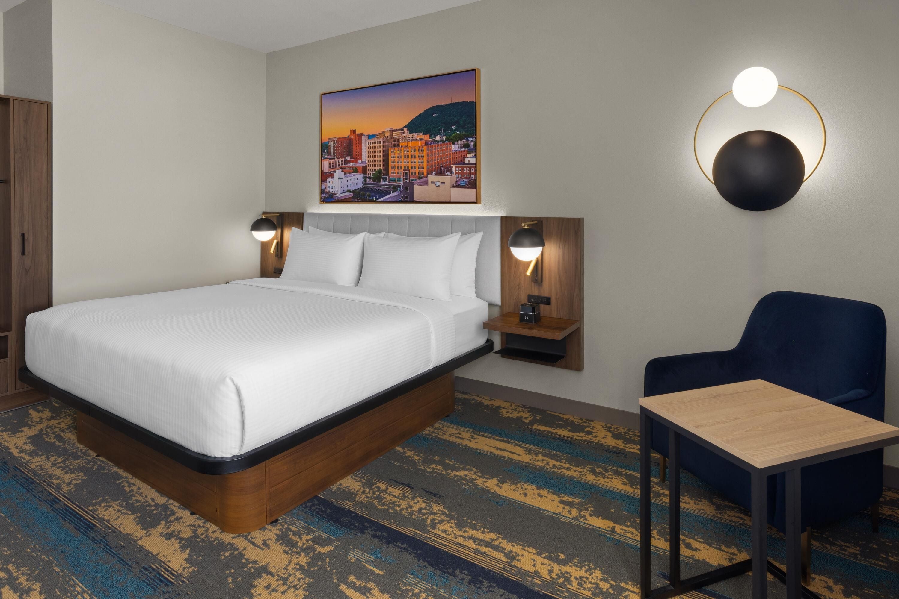 Quarto de hotel com uma cama grande, uma escrivaninha, duas luminárias de cabeceira e uma pintura da paisagem urbana na parede.