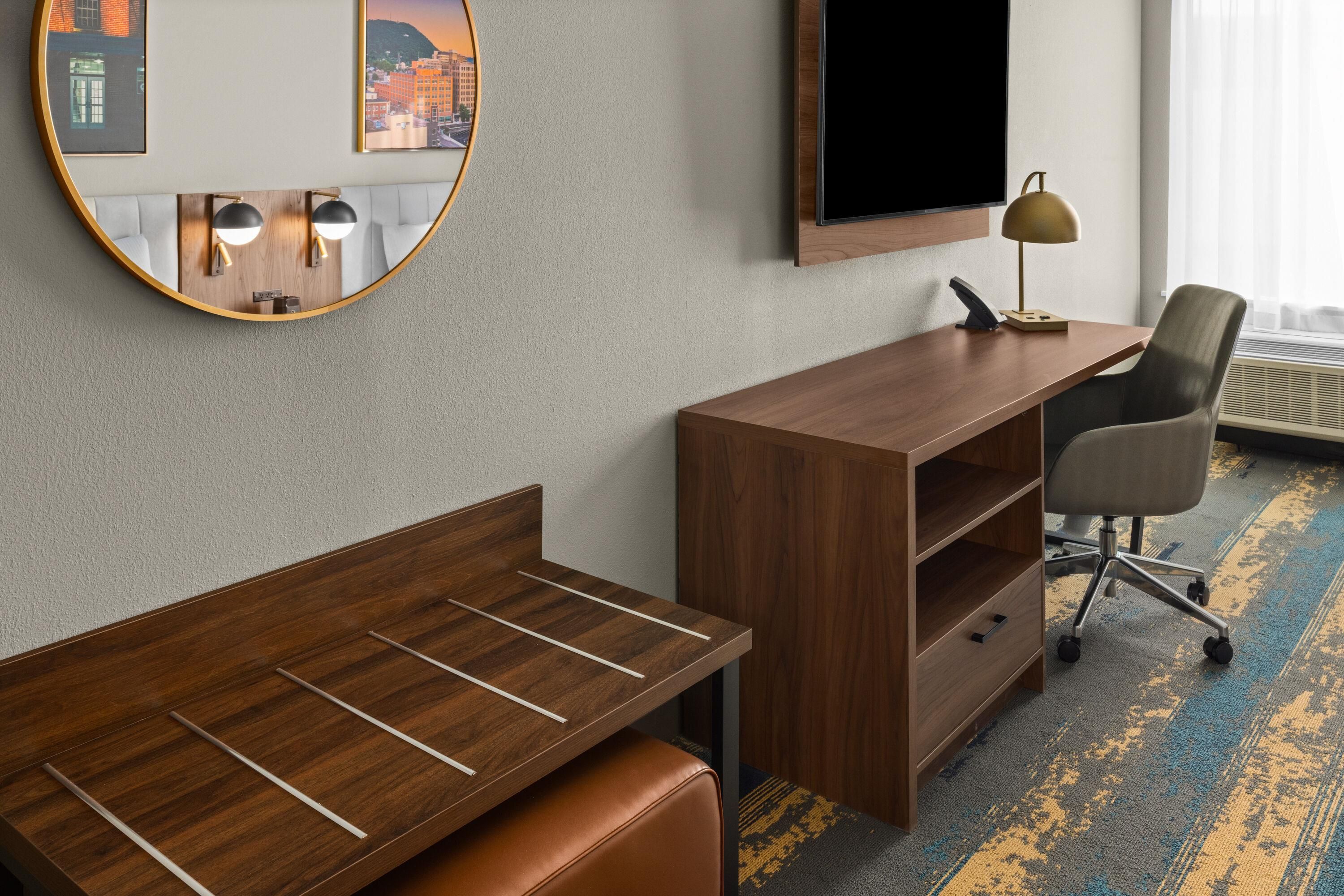 A hotel room with a wooden desk, a chair, a television mounted on the wall, and a round mirror.