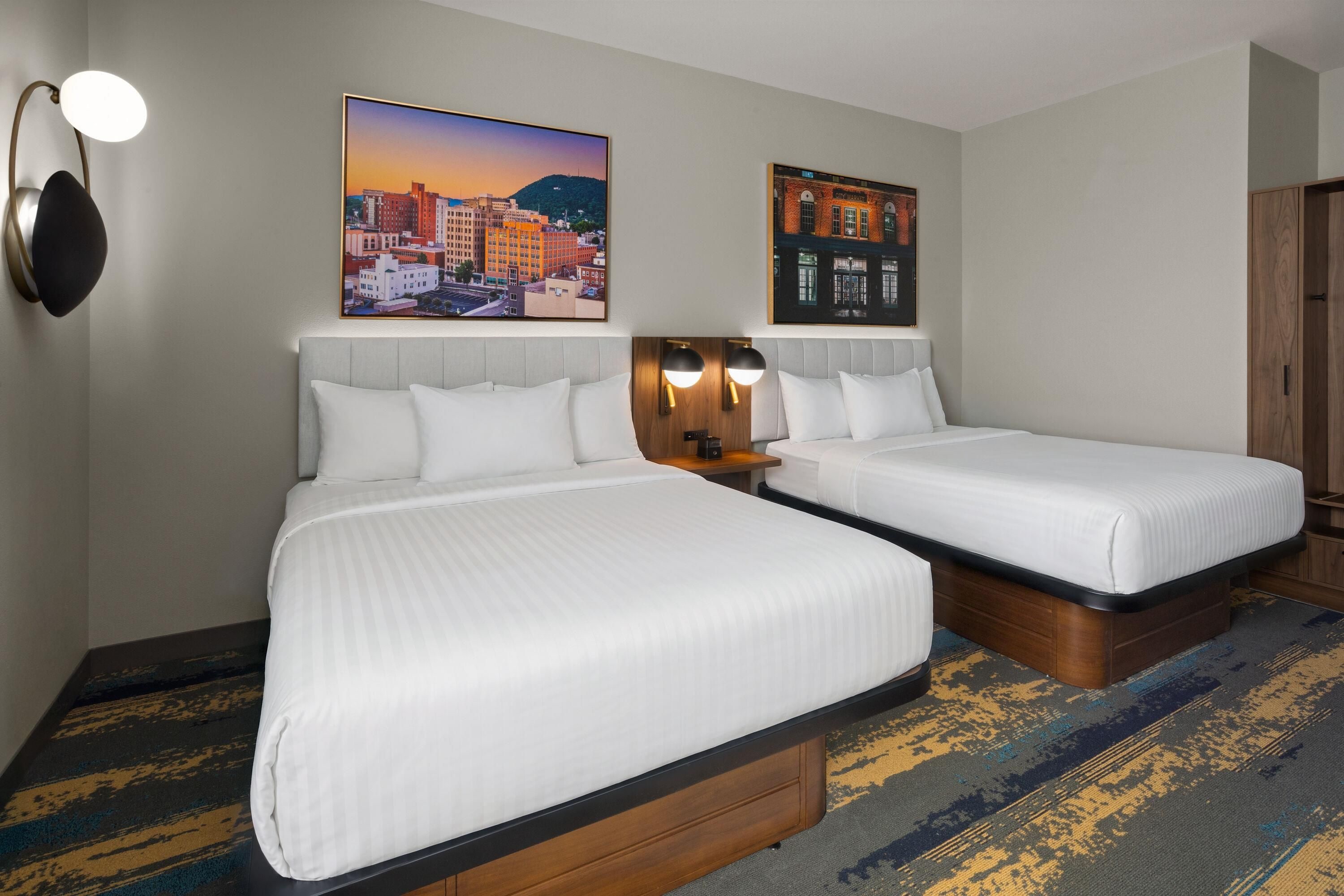 A hotel room with two beds, a cityscape painting above each bed, a nightstand with a lamp, and a patterned carpet.