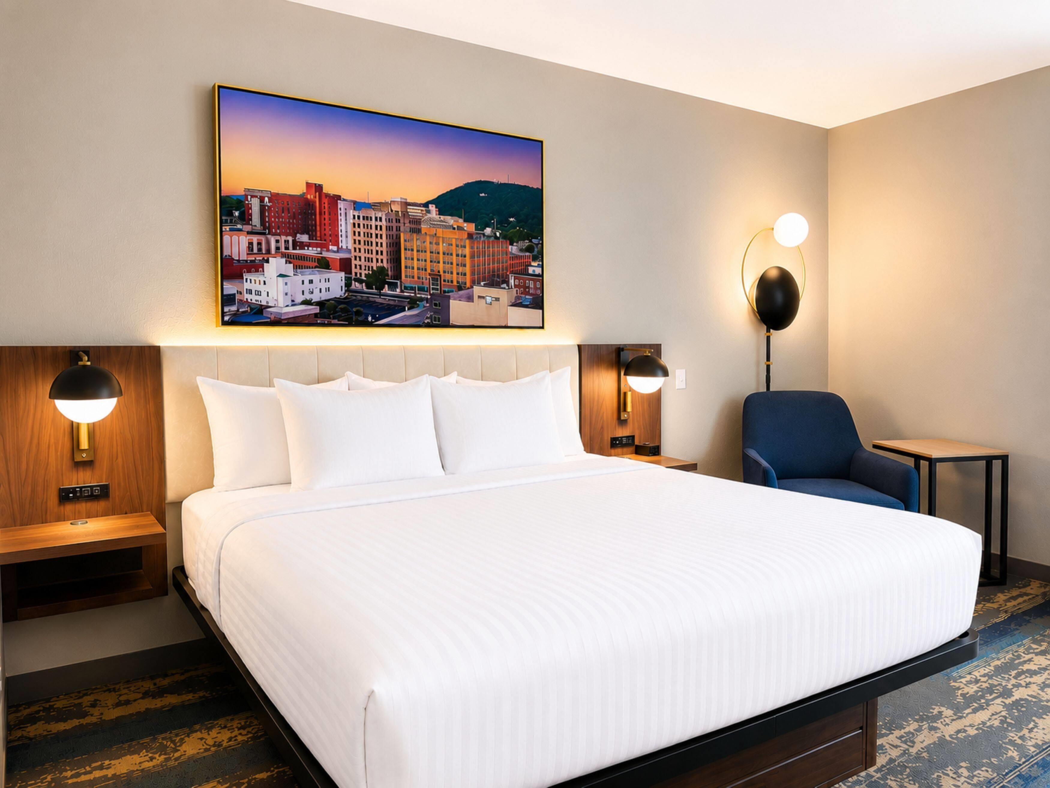 A hotel room with a large bed, two bedside tables, a chair, a desk, and a cityscape painting on the wall.