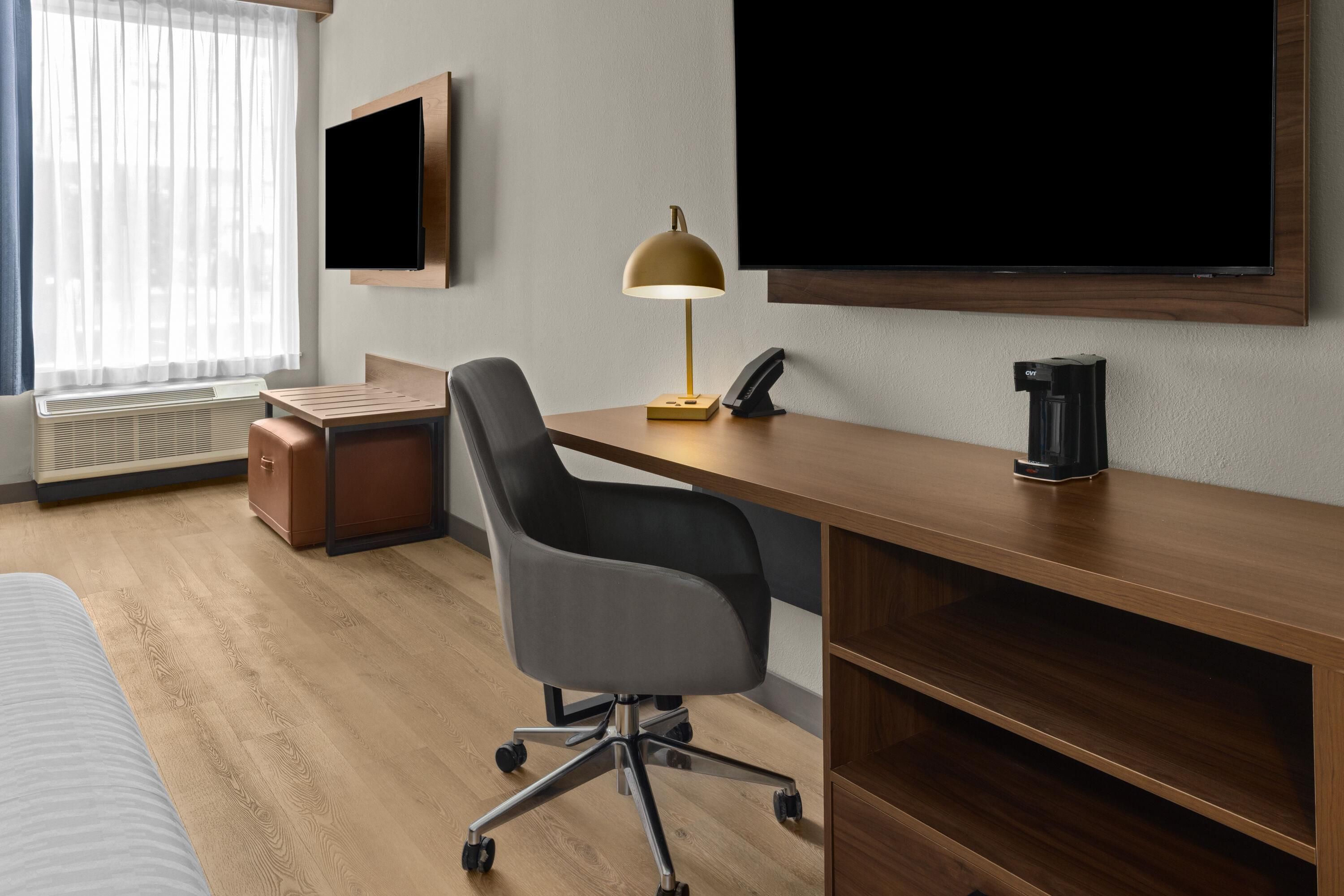 Ein Hotelzimmer mit einem Holztisch, einem Stuhl, einem Flachbildfernseher, einer Lampe, einem Heizkörper und einem Bett.