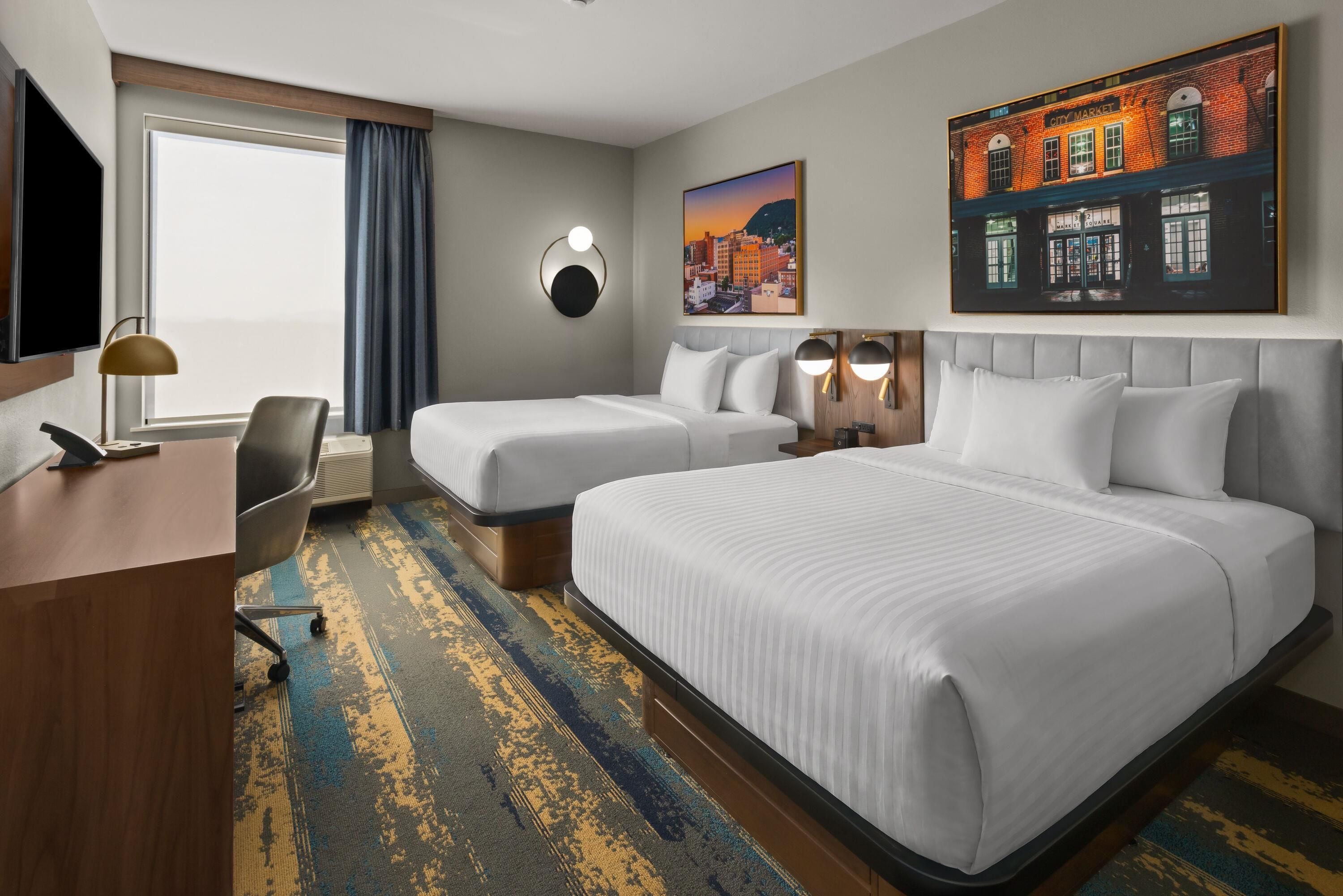 A hotel room with two beds, a desk, a chair, a TV, and a large window with curtains.