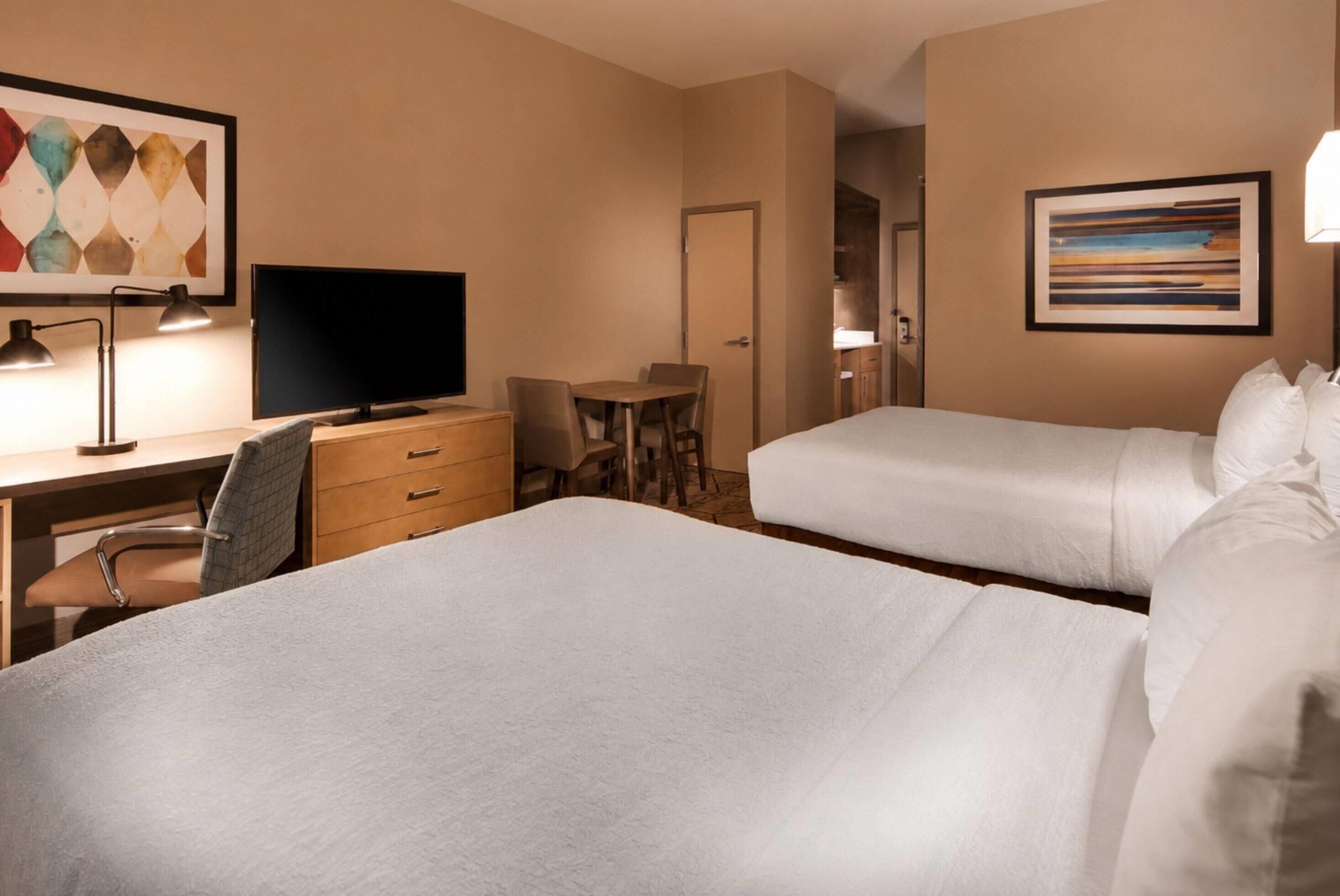 A hotel room with two beds, a desk with a chair, a television, and artwork on the walls.