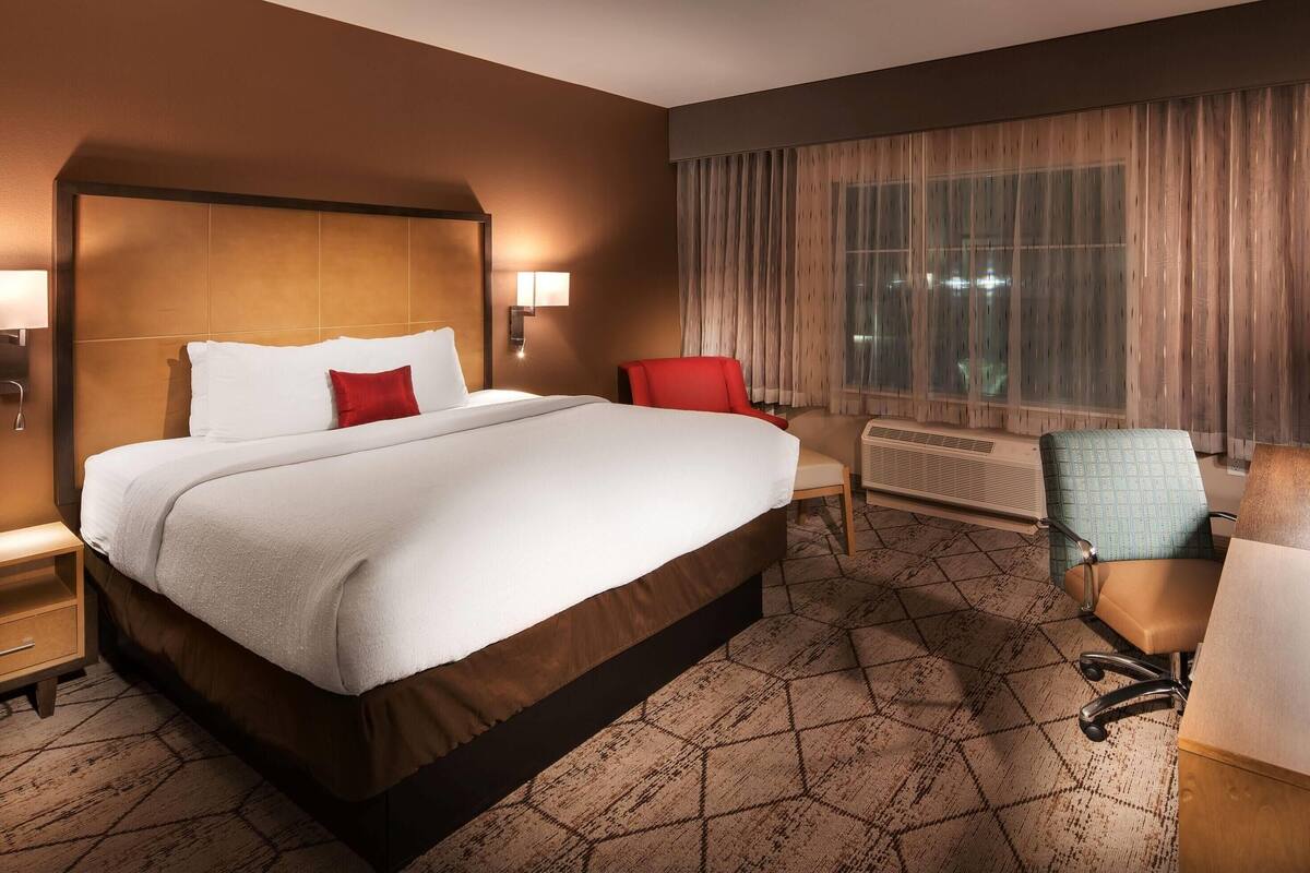 A hotel room with a large bed, a desk, a chair, a nightstand, and a window with curtains.