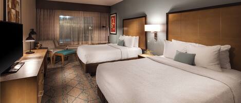 Quarto de hotel com duas camas, uma escrivaninha, uma cadeira, uma televisão e uma janela com cortinas.