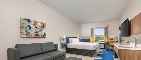 A modern hotel room with a gray sofa, a small table, a bed with a blue headboard, a desk with a chair, a large window with curtains, and a colorful abstract painting on the wall.