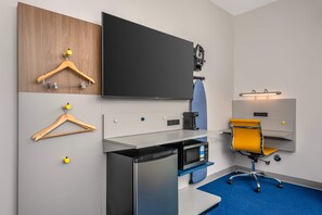 Hotelværelse med fladskærms-TV, kaffemaskine, mikroovn, skrivebord, stol og blåt tæppe.
