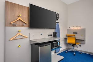 Quarto de hotel com televisão de tela plana, cafeteira, micro-ondas, escrivaninha, cadeira e tapete azul.
