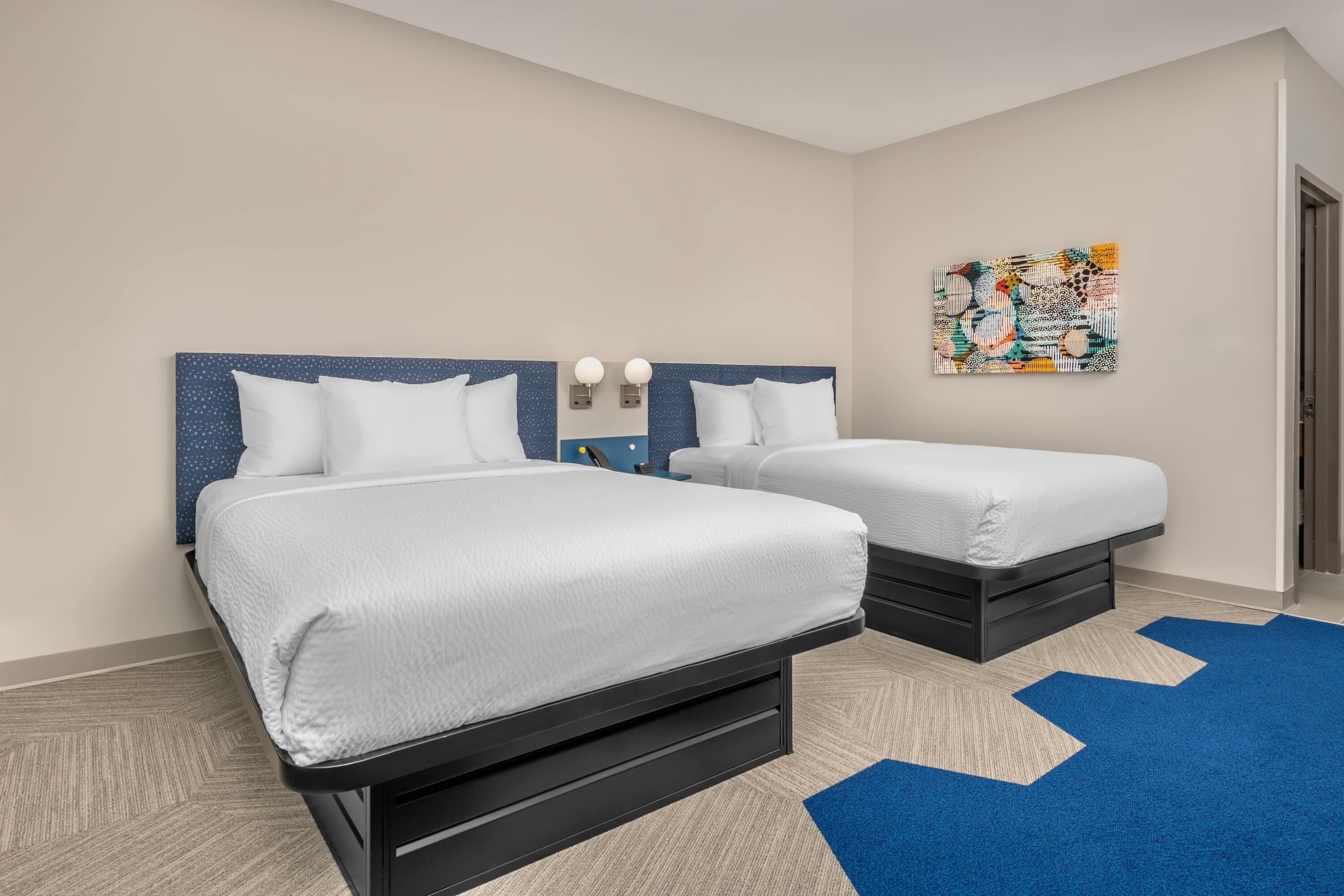A hotel room with two beds, a blue and white patterned wall, and a colorful abstract painting.