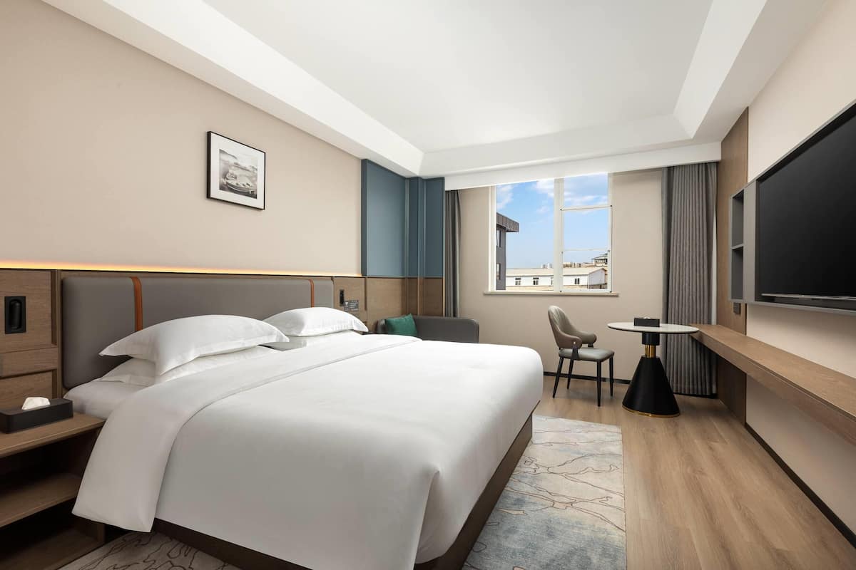 Una habitación de hotel moderna con una cama grande, un escritorio, una silla, un televisor y un ventanal con vista a la ciudad.