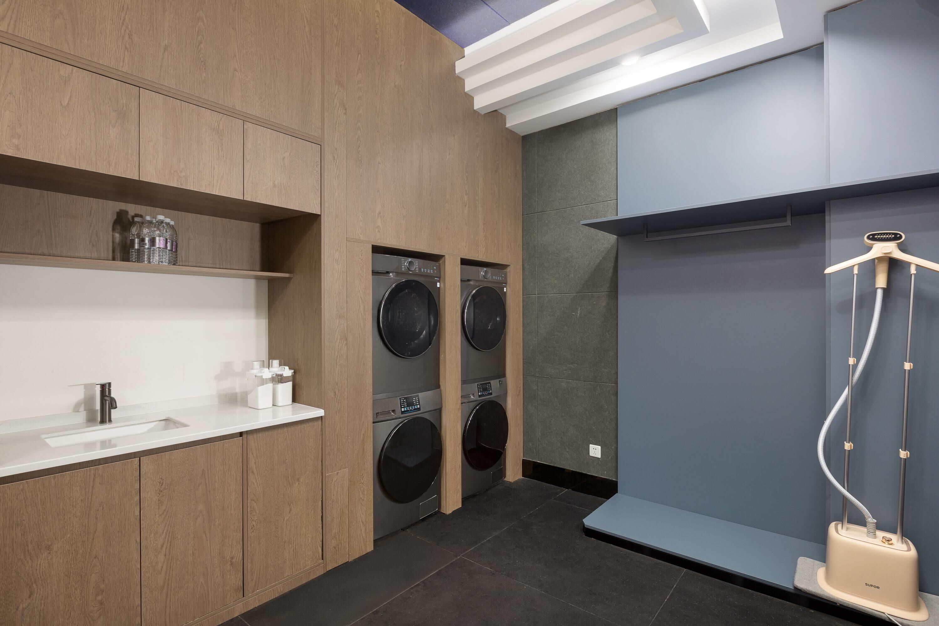 A modern bathroom with wooden cabinetry, a sink, two wall-mounted washing machines, and a shower unit.