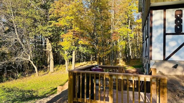 Une terrasse en bois avec un barbecue, entourée d’arbres et bordée d’un bâtiment à la façade blanche et aux éléments décoratifs sombres.