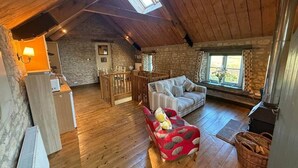 Un acogedor cuarto estilo loft con techo de madera, paredes de piedra, chimenea, un sofá, un reposapiés rojo y un comedor.