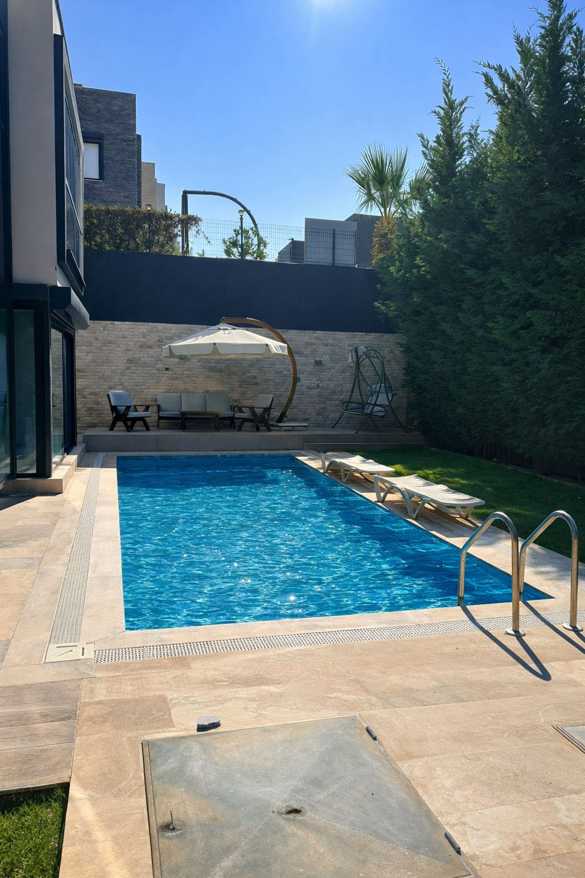 Ein Swimmingpool mit Liegestühlen und Sonnenschirm, umgeben von einer Steinmauer und viel Grün.