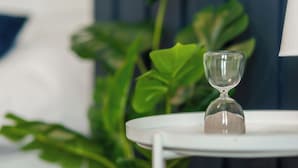 Een glazen zandloper op een witte tafel, met een groene plant op de achtergrond.
