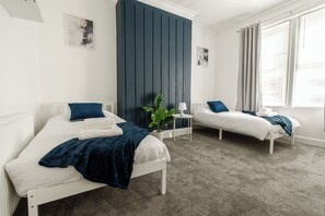 Quarto com duas camas, uma parede de destaque escura, uma mesinha e uma janela com cortinas.