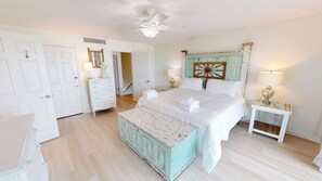 Une chambre à coucher comprenant un lit, une commode, une table de chevet, un ventilateur de plafond et une porte.