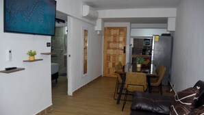 Moderni olohuone, jossa on nahkasohva, ruokailutila tuoleineen, keittiö jääkaapilla sekä seinälle asennettu televisio.