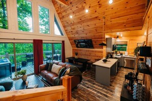 A cabin interior with a living area, dining space, kitchen, and a deck with a view of the forest. - Back Yard Cliff View | Elevated Cabin | Hot Tub | Fire pit (Lee County)
