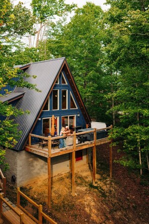 A cabin with a steep gabled roof, a deck with outdoor seating, and a surrounding forest. - Back Yard Cliff View | Elevated Cabin | Hot Tub | Fire pit (Lee County)