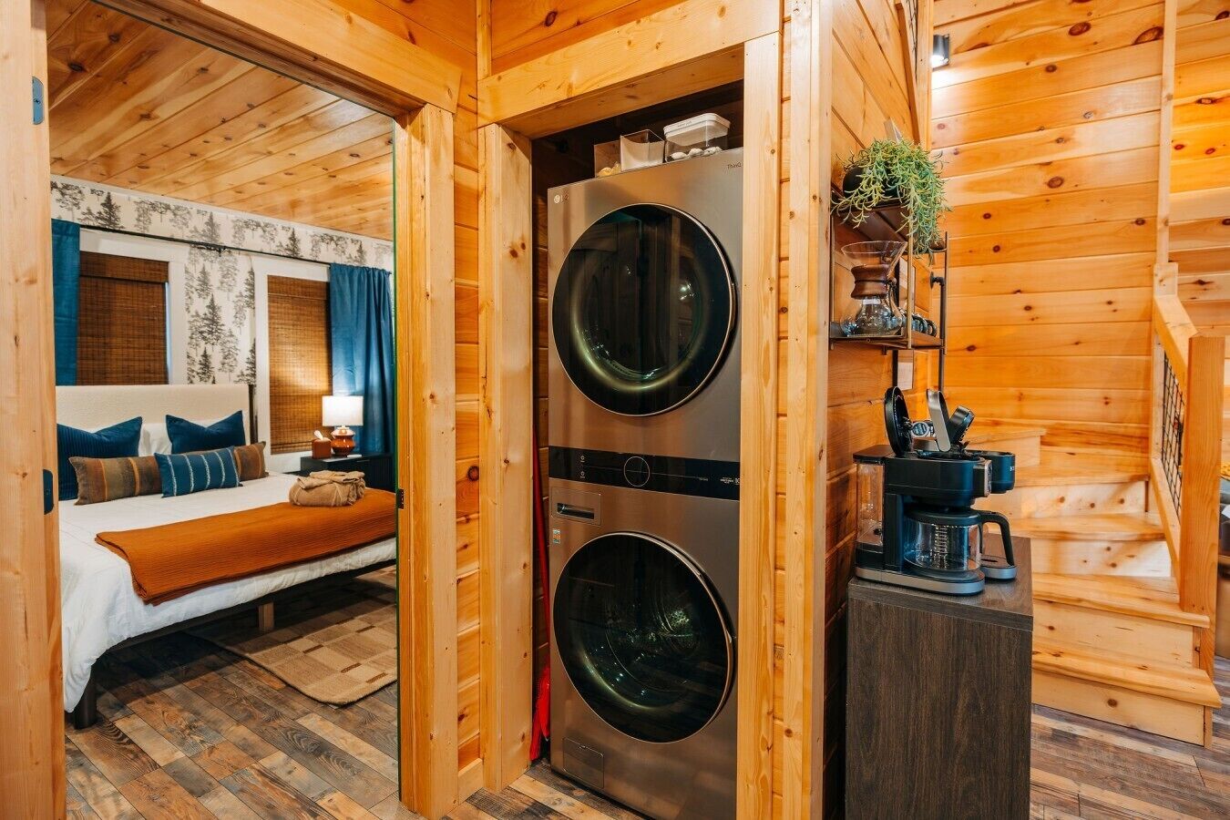 A wooden cabin interior with a bed, two washing machines, a coffee maker, and a shelf with a plant.