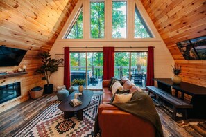 A cozy wooden interior with a fireplace, a sofa, a coffee table, and a large window overlooking a deck. - Back Yard Cliff View | Elevated Cabin | Hot Tub | Fire pit (Lee County)