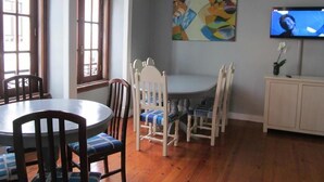 A dining area with a round table, wooden chairs, a television on the wall, and a painting. - Casa Azul Hostel (Sintra)