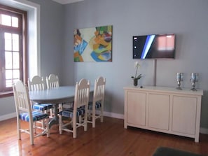 A dining area with a round table, six chairs, a cabinet, a TV, and a painting on the wall. - Casa Azul Hostel (Sintra)