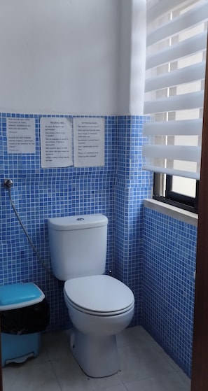Un baño pequeño con inodoro, bidé y paredes de azulejos azules. Hay carteles con normas del baño y una nota sobre no arrojar toallas de papel al inodoro.