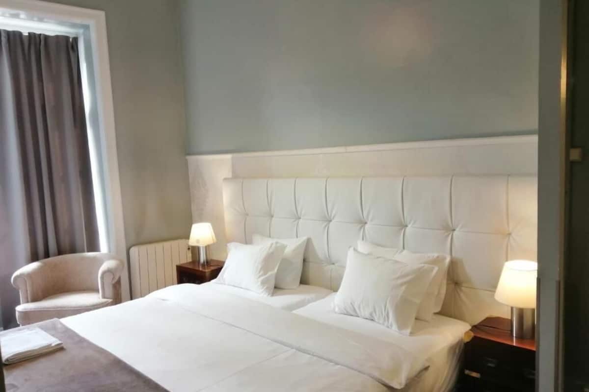 Habitación de hotel con una cama grande, dos mesitas de noche con lámparas, una silla y un ventanal con cortinas.