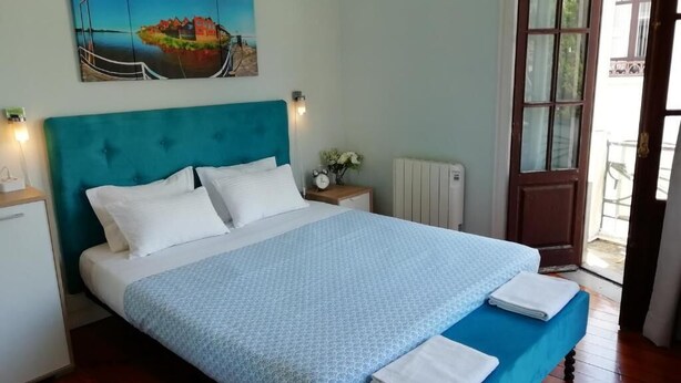 A bedroom with a bed, a nightstand, a painting, a window, and a door. - Casa Azul Hostel (Sintra)