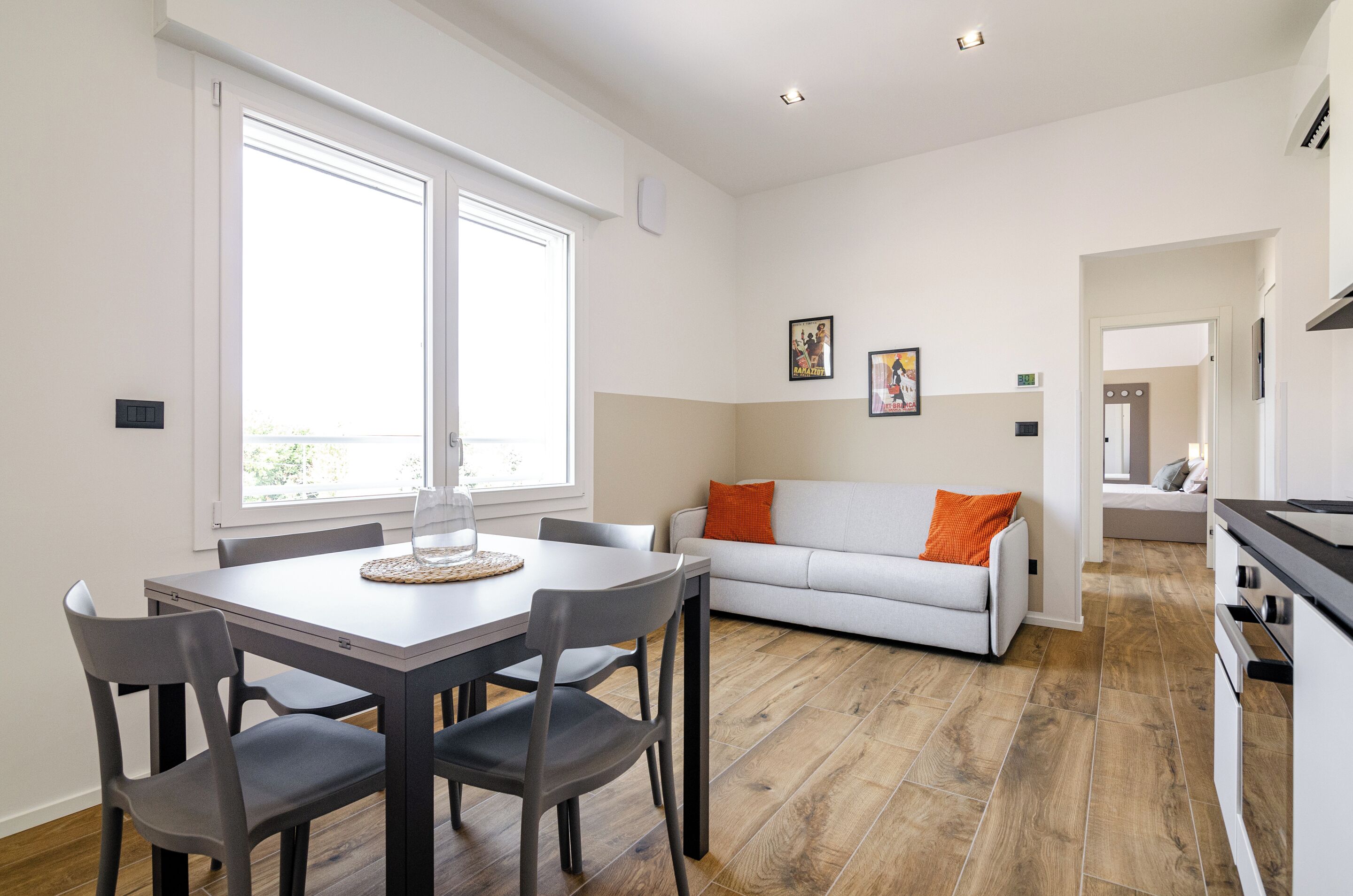 A modern kitchen with a dining area, a sofa with orange pillows, and a view through a window.