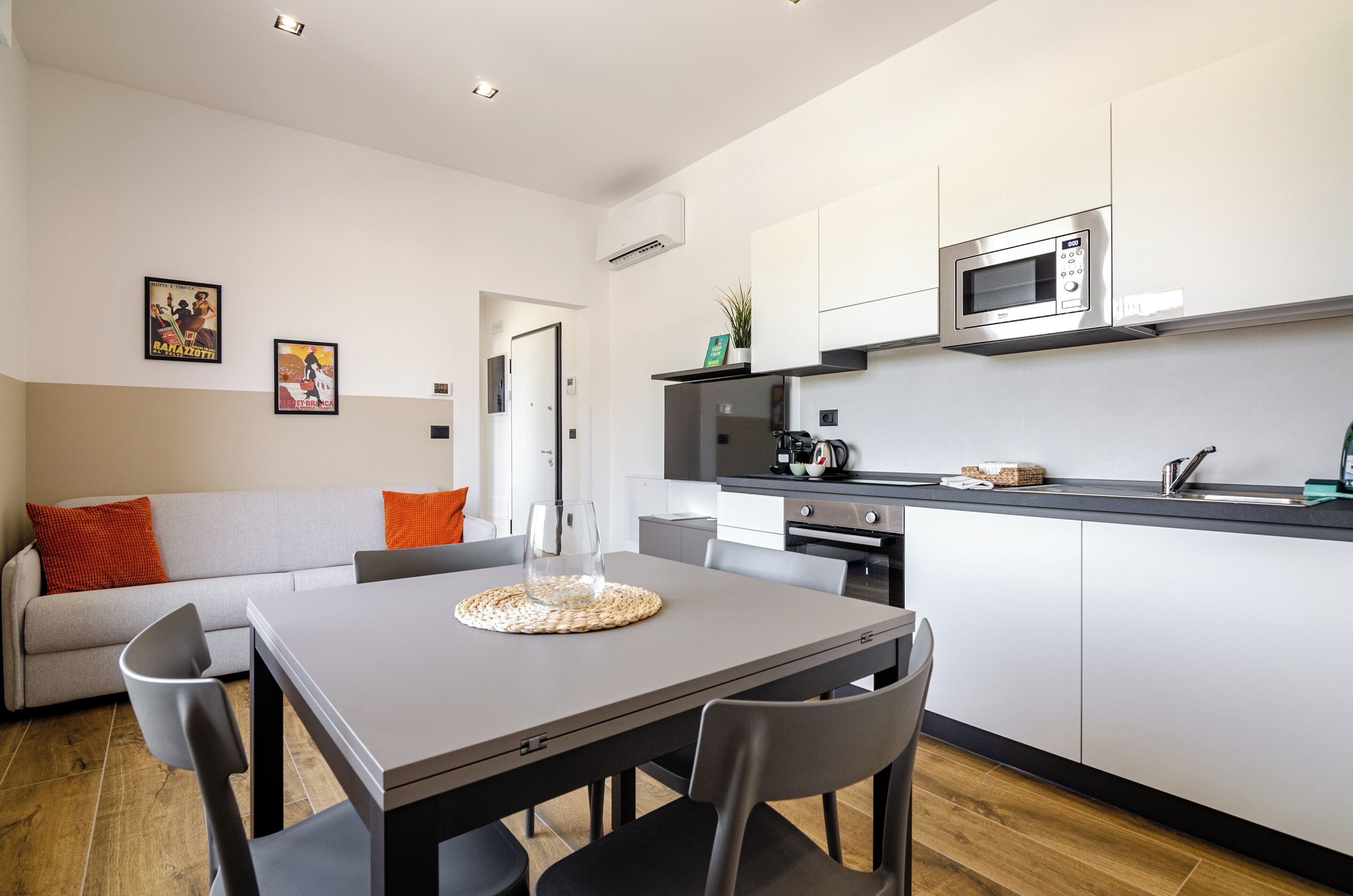 A modern kitchen with a microwave, stove, and sink. There is a dining table with chairs and a sofa with orange cushions.