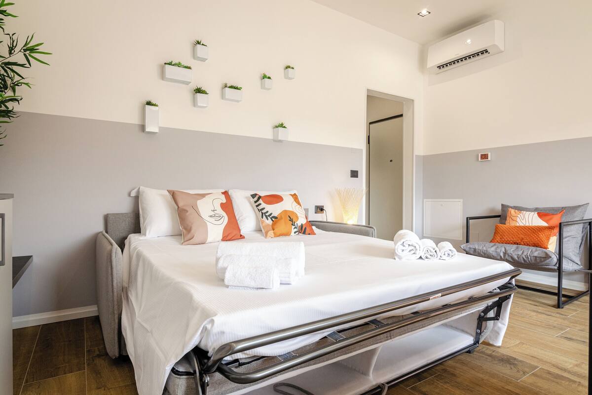 A hotel room with a bed, a chair, a wall-mounted air conditioner, and several small potted plants.