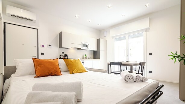 A modern hotel room with a bed, two pillows, a desk with a chair, a window, and a kitchen area.
