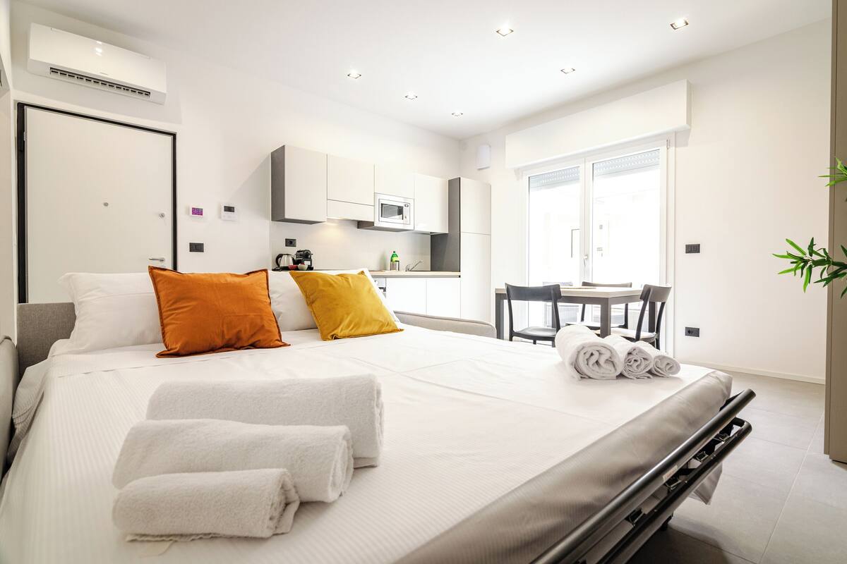 A modern hotel room with a bed, two pillows, a desk with a chair, a window, and a kitchen area.